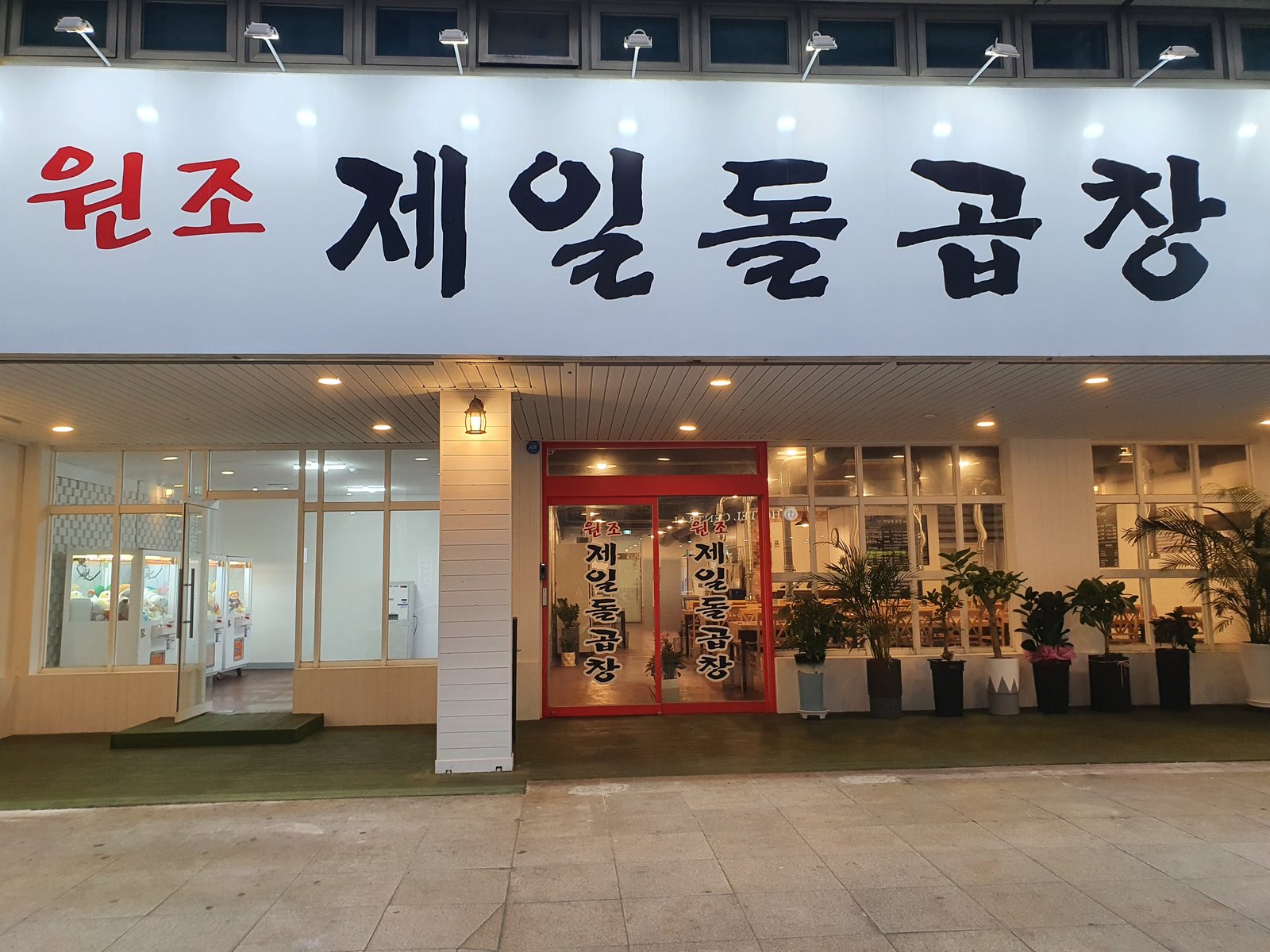 광주 서구 상무연하로 67, 1965원조제일돌곱창 상무점 외관. 흰 간판에 붉은 ‘원조’와 검은 붓글씨 ‘제일돌곱창’이 적혀 있고, 붉은 문틀 유리문 옆으로 화분이 줄지어 있다
