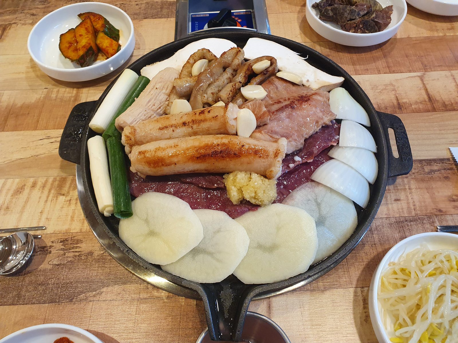 제일돌곱창 상무점의 한우소금모듬구이 — 둥근 판에 대창·곱창·염통·특양과 대파, 양파, 새송이, 감자, 통마늘을 둘러 담았고 오이무침·콩나물무침이 곁들여 있다